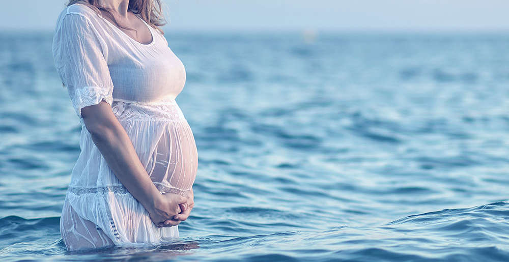 Mujer embarazada pasea dentro del mar con vestido blanco tocando su barriga